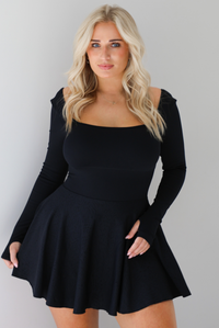 Woman wearing a black athleisure dress that has long sleeves with thumbhole cutouts, a scooped neckline, fitted bodice, and flowy skirt. The photo shows a closeup of the dress, where you can see the bodice has a ribbed texture, and the skirt has a squiggly line texture. The photo is taken indoors against a white wall.