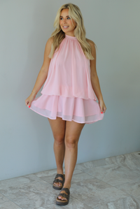 Woman wearing a pink dress that has a halter tie neckline, a cinched detail at the hips, and a flowy design indoors against a white wall with white marble flooring. 