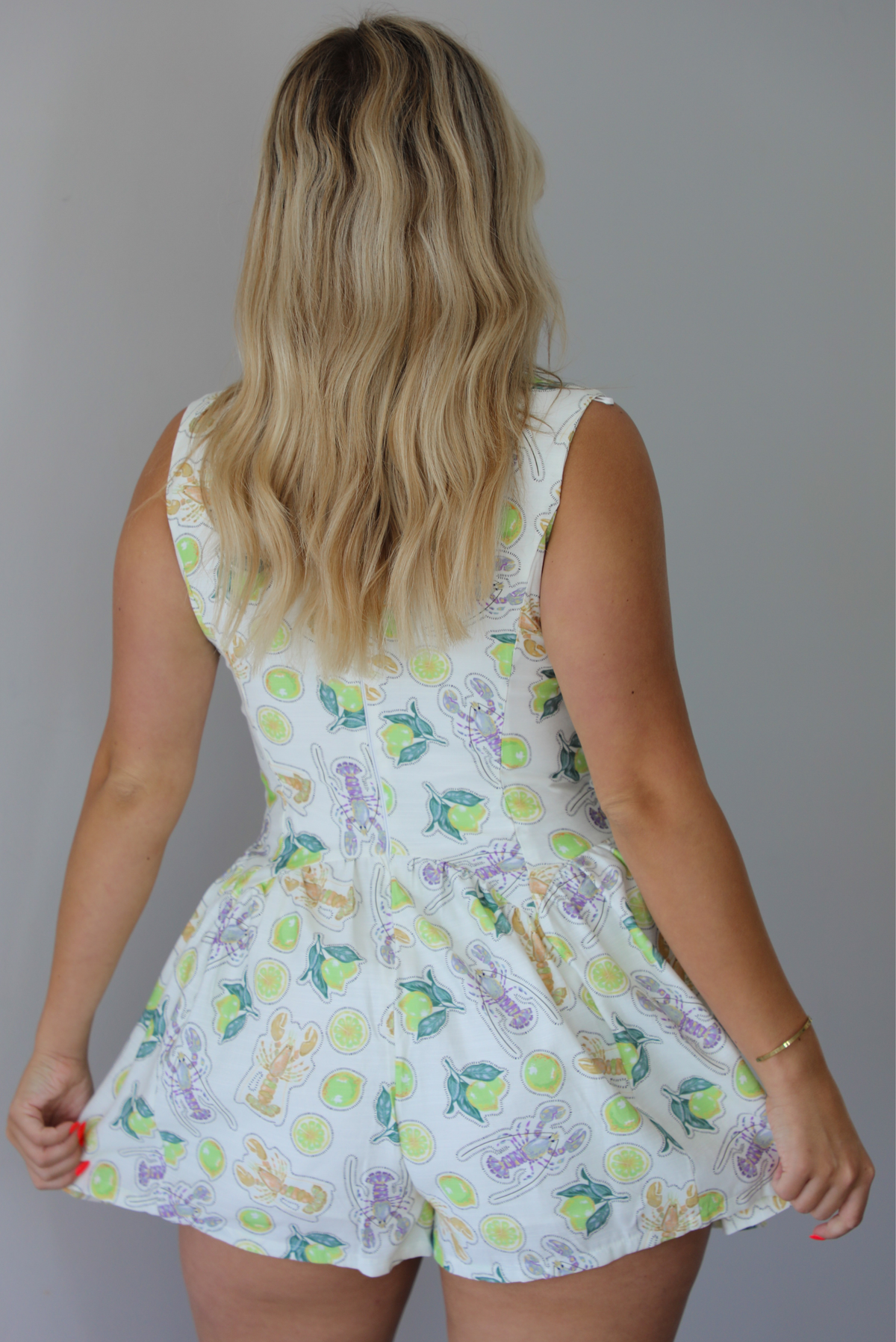 Woman wearing a white and multi-colored romper that has a lime and lobster pattern throughout, a plunging v-neckline, cut out and tie details at the bodice and waist, and slightly flowy shorts indoors against a white wall. The photo shows the back of the romper. 