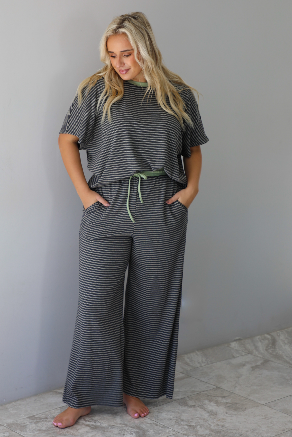 A woman wearing a charcoal and white striped two piece matching set with green trim detailing at the neckline and waistline, featuring an oversized short sleeved top and high waisted pants, standing indoors against a white wall with white marble flooring. 