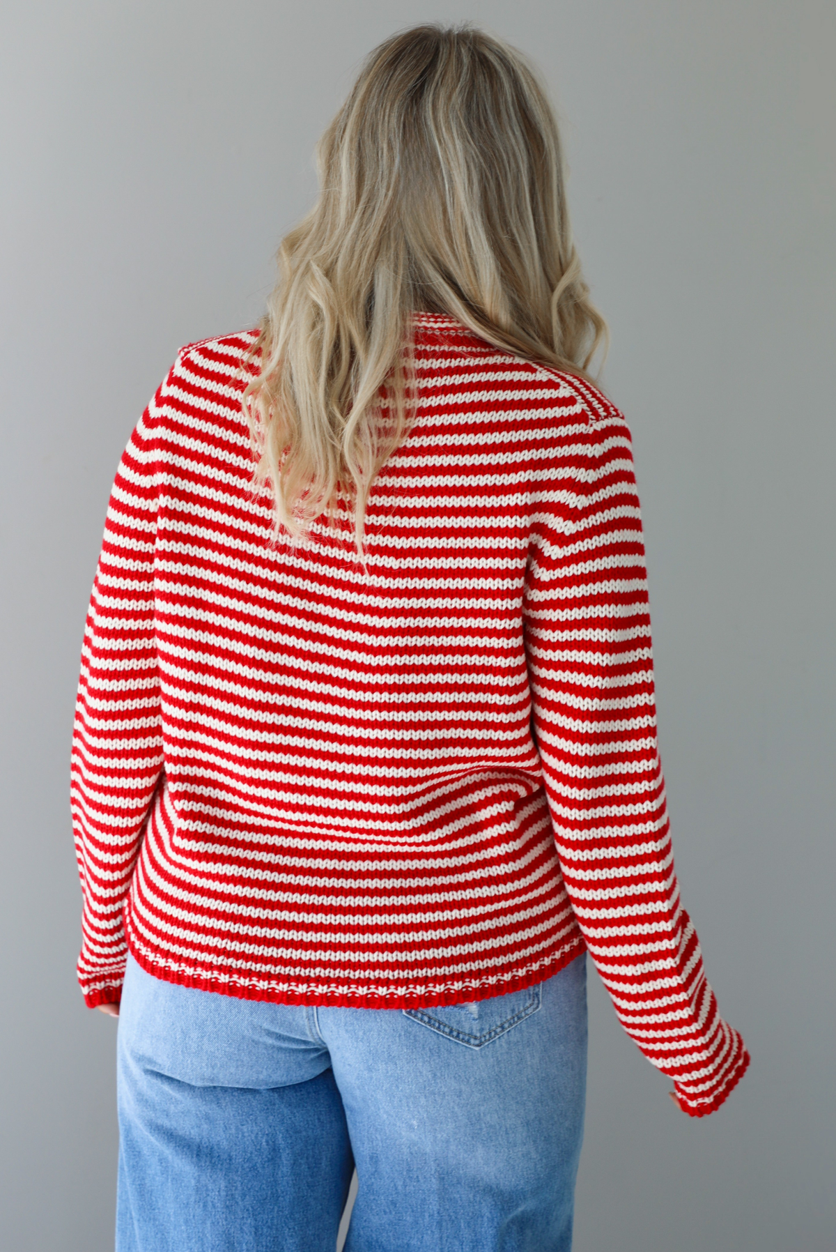 Woman wearing a red and white striped sweater with high waisted medium wash flared hem denim. The photo shows a closeup of the back of the sweater. The photo is taken in front of a white wall.