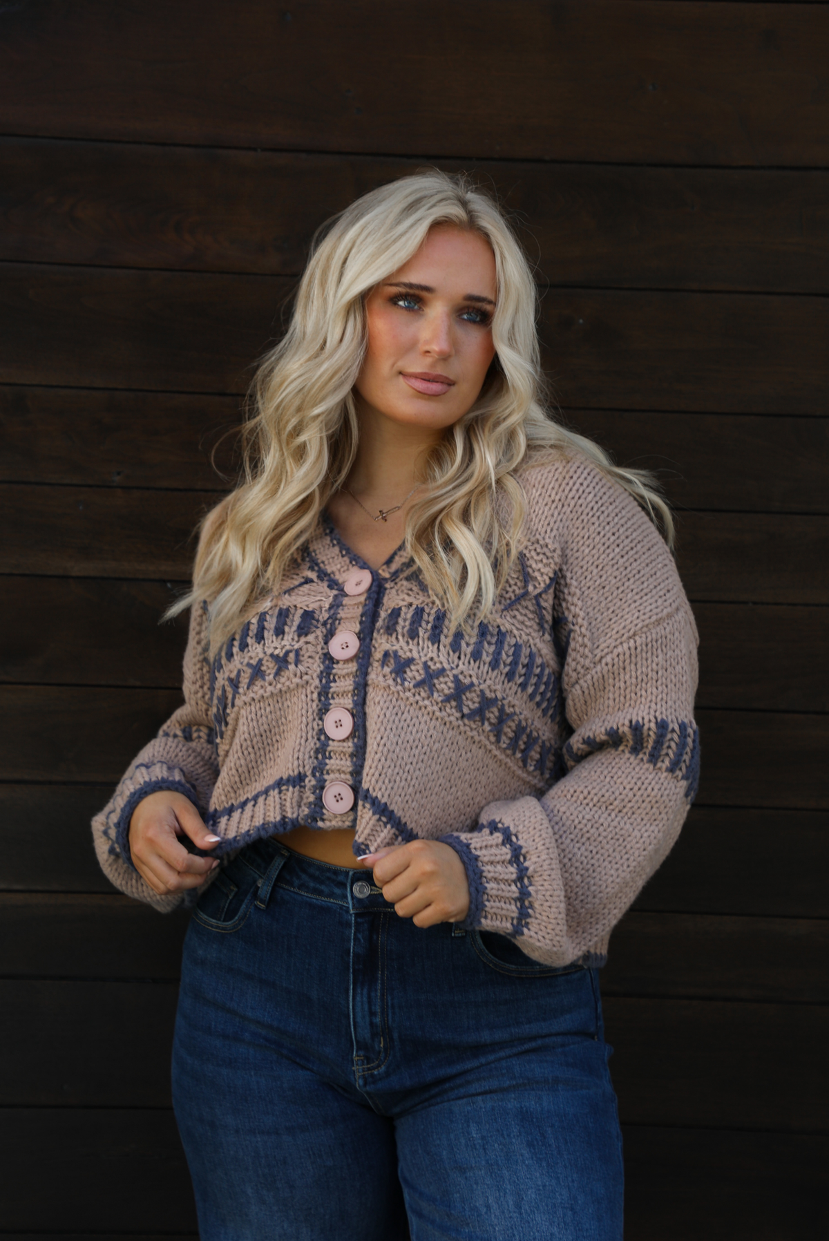 Woman model wearing a mocha brown cardigan that has grey stitching details throughout. The cardigan has a cropped cut, is oversized, and has functional button closures in the front. She's wearing the cardigan with dark denim jeans. The photo showcases a close up of the cardigan. The photo is taken against a dark brown wooden door outdoors. 