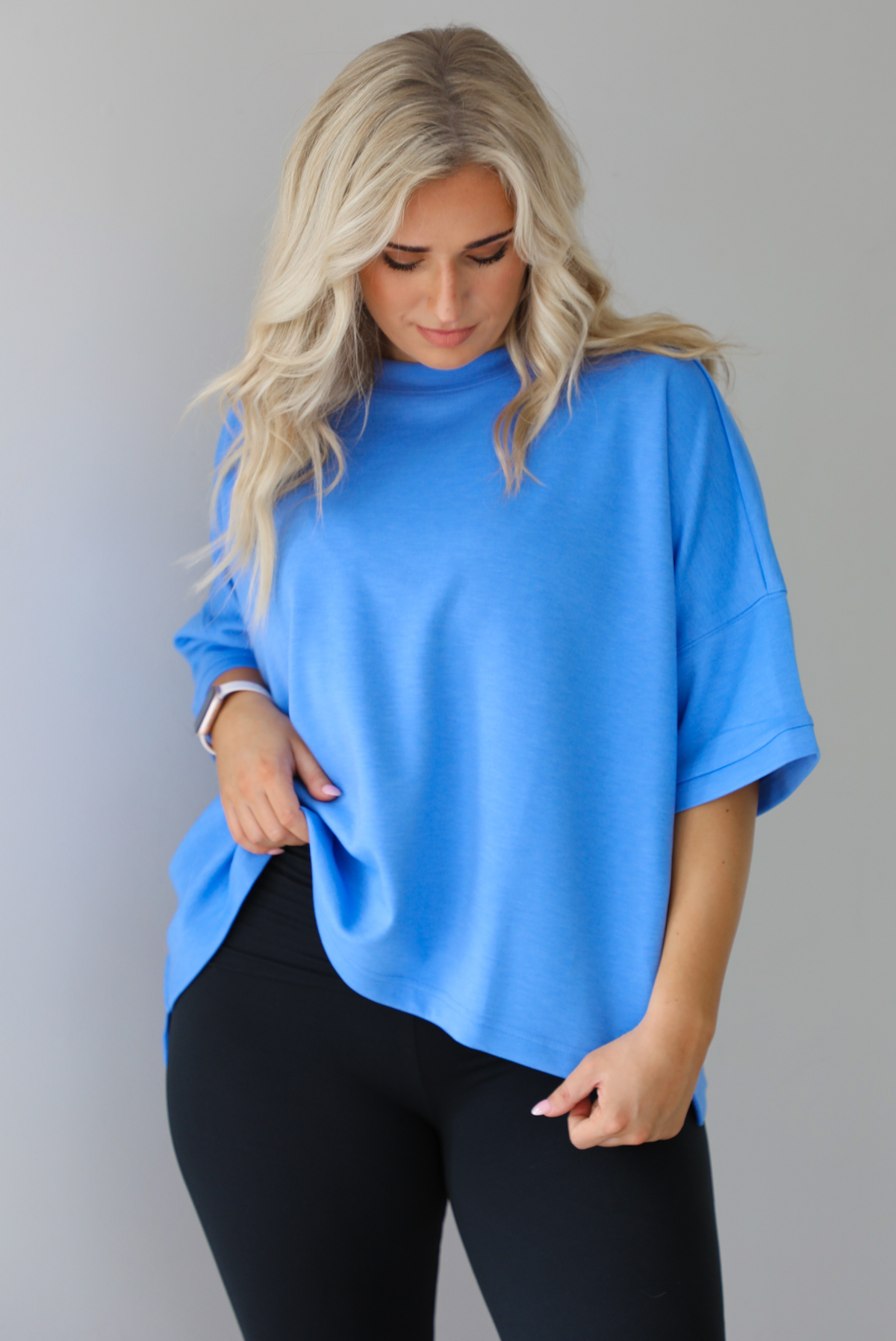 Woman wearing an oversized blue box tee with black leggings. The photo shows a closeup of the tee. The photo is taken indoors against a white wall with white marble flooring. 