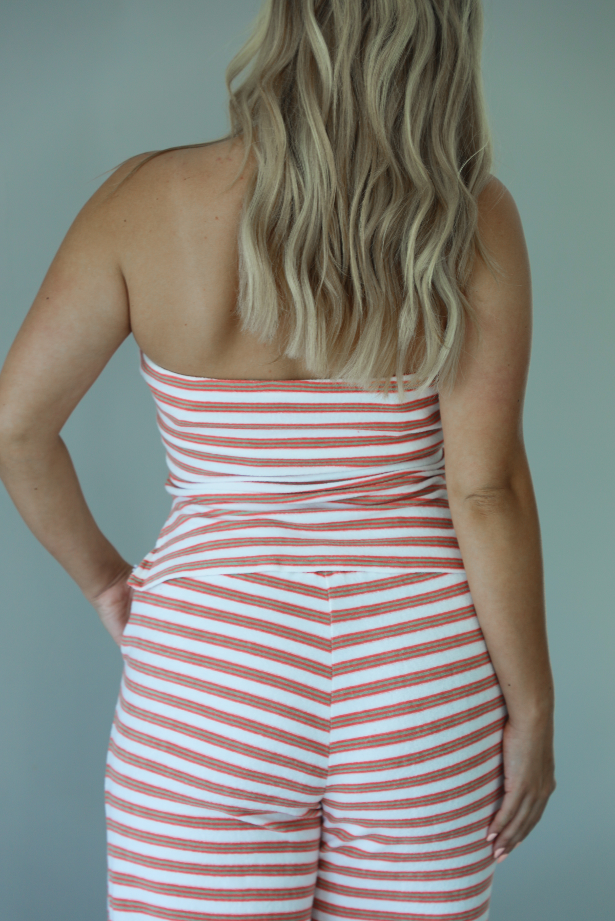 Woman wearing an orange, white, and green striped pattern set that has a strapless tube top and high waisted pants indoors against a white wall and shows a closeup of the back of the top. 