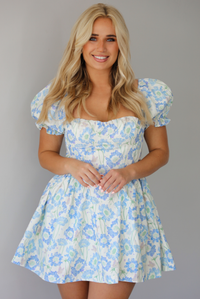 Woman wearing a white, blue, and green floral print mini dress that has short, puffed sleeves, a fitted corset bodice, and a flowy skirt indoors against a white wall.