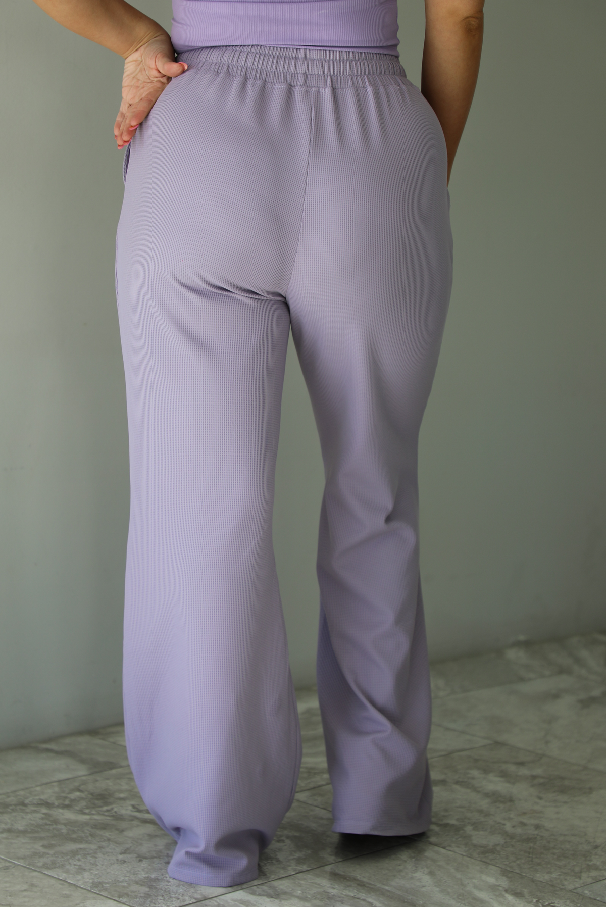 Woman wearing a two piece lavender set featuring a strapless, fitted tube top, and high waisted, flared hem pants. The photo is taken indoors against a white wall and shows a closeup of the back of the pants, which are high waisted with an elastic and drawstring waistline, with functional pockets on either side, has a repeating squared textured design, and a flared hem.