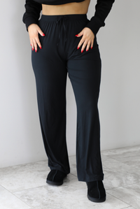 The photo features a model wearing high waisted black pants that have an elastic and drawstring waistline, a slightly flared hem, and are super soft and stretchy. They are worn with black ugg boots. The photo is taken indoors against a white wall with white marbled flooring. 