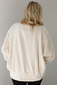 Woman wearing a sherpa sand oversized sweater with a fitted black jumpsuit underneath. The photo shows a closeup of the back of the sweater and the raw seam details. The photo is taken indoors against a white wall with white marble flooring. 