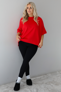 Woman wearing an oversized red t-shirt with black, fitted lettings, scrunchie socks, and black ugg boots. The photo is taken indoors against a white wall with white marbled flooring. 