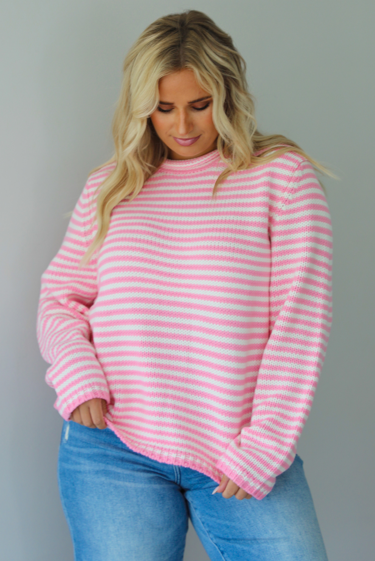 A woman wearing a pink and white striped sweater paired with light wash denim jeans, standing indoors against a white wall.