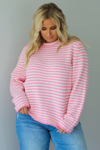 A woman wearing a pink and white striped sweater paired with light wash denim jeans, standing indoors against a white wall.