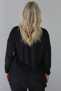Woman wearing a black two piece set that features an oversized long sleeved top and fitted leggings. The photo shows a closeup of the back of the top. The photo is taken indoors against a white wall.