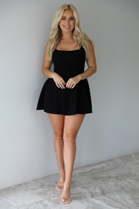 Woman wearing a black mini dress with thick straps, a fitted bodice, and flowy skirt indoors against a white wall with white marble flooring. 