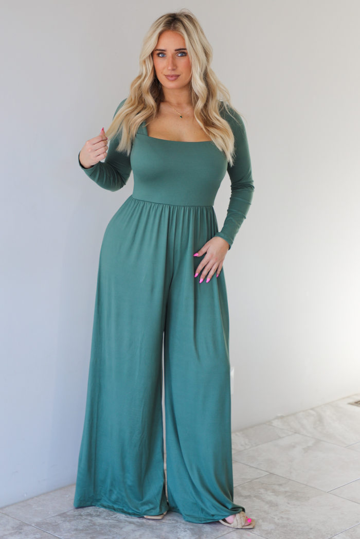 Woman wearing a green jumpsuit that has a shared neckline, long sleeved, and long, flowy pants.  The photo is taken indoors against a white wall with white marble flooring. 