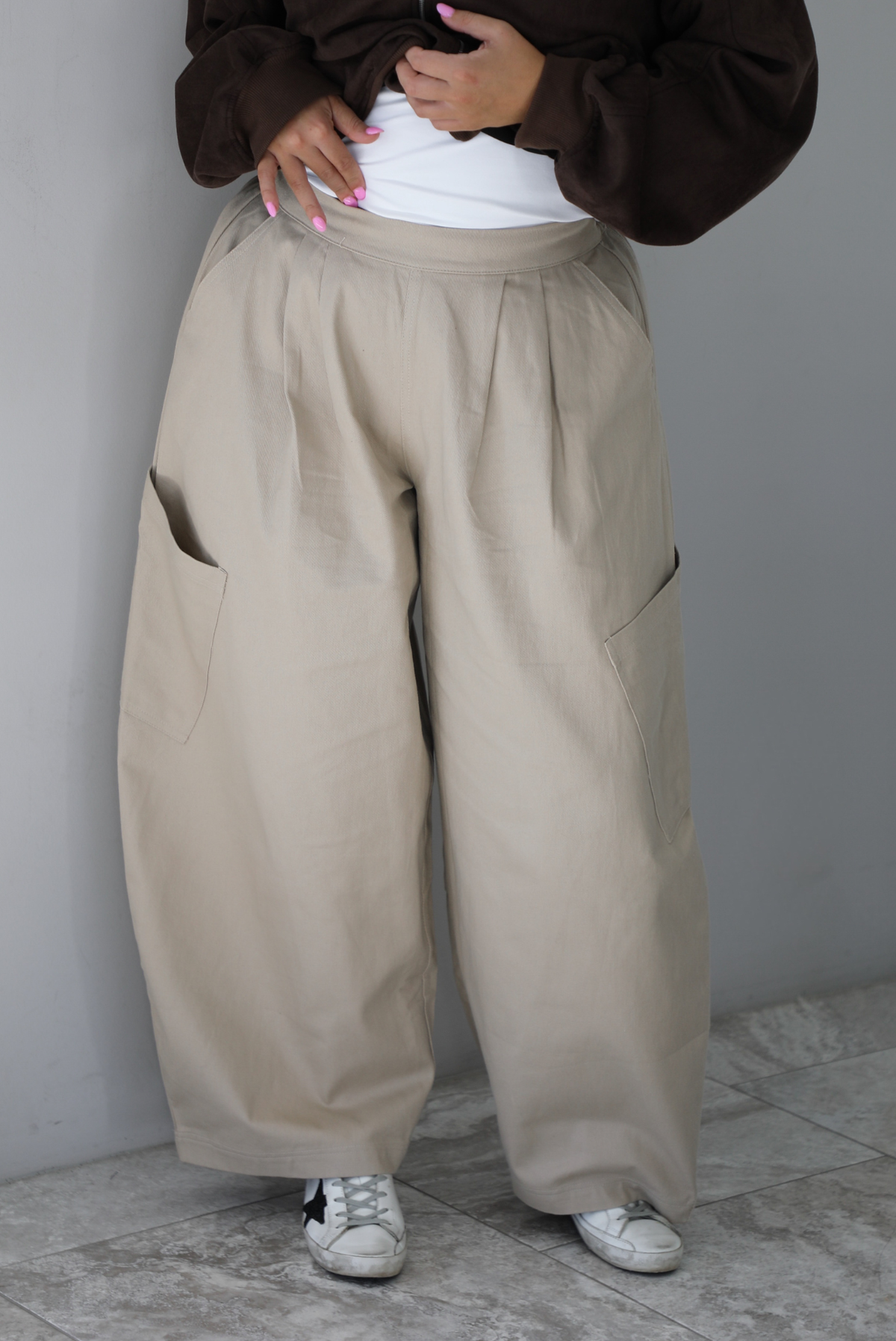A woman shown from the waist down wearing a pair of sand baggy pants with functional pockets on either side and more pockets further down on the thigh just above the knees, standing indoors against a white wall with white marble flooring. 