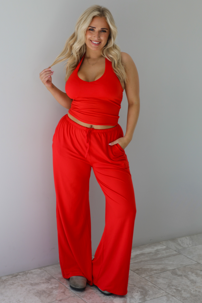 Woman wearing a red two piece set featuring a  tank top and slouchy pants. The photo is taken indoors against a white wall with white marble flooring. 
