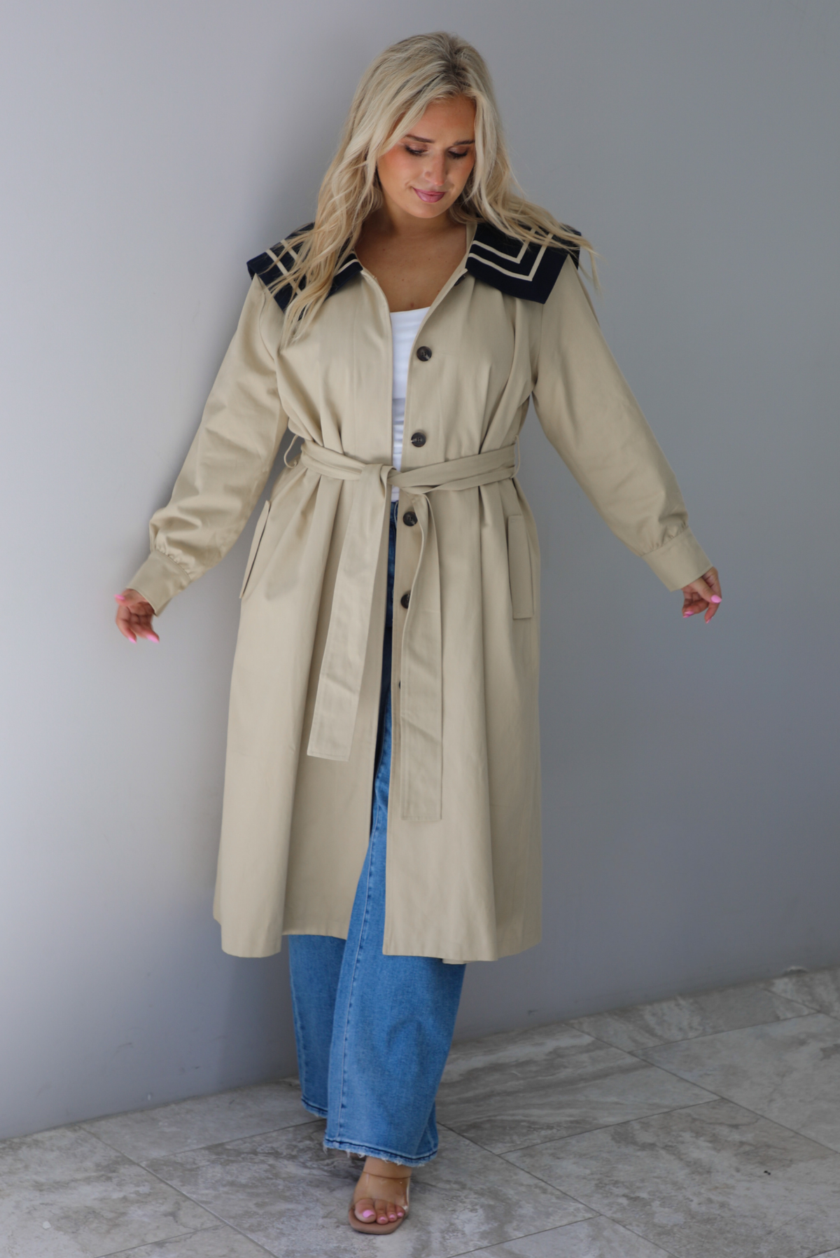 A woman wearing a tan trench coat with a navy collared neckline, functional button closures in the front, functional pockets on either side, with a tie waistline, paired with light wash flared hem denim jeans, standing indoors against a white wall with with white marble flooring. 