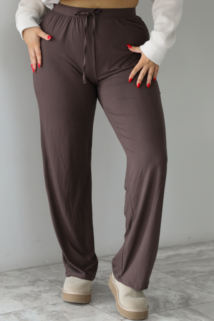 The photo features a model wearing high waisted brown pants that have an elastic and drawstring waistline, a slightly flared hem, and are super soft and stretchy. They are worn with beige ugg boots. The photo is taken indoors against a white wall with white marbled flooring. 