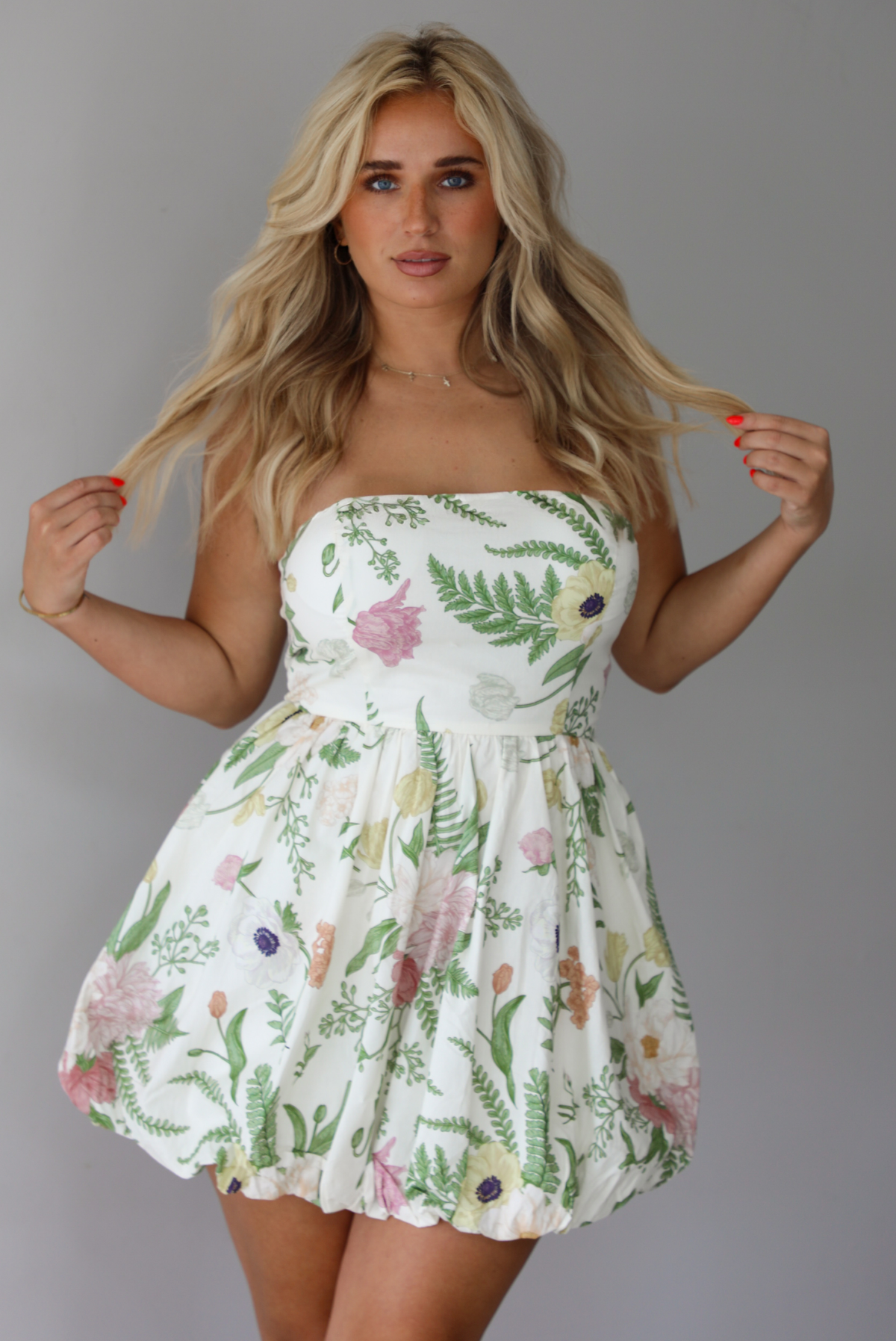 Woman wearing a cream strapless mini dress that has a multi-colored floral pattern throughout indoors against a white wall.