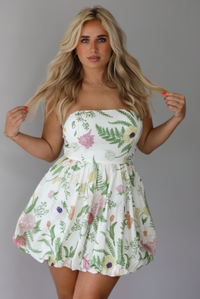Woman wearing a cream strapless mini dress that has a multi-colored floral pattern throughout indoors against a white wall.
