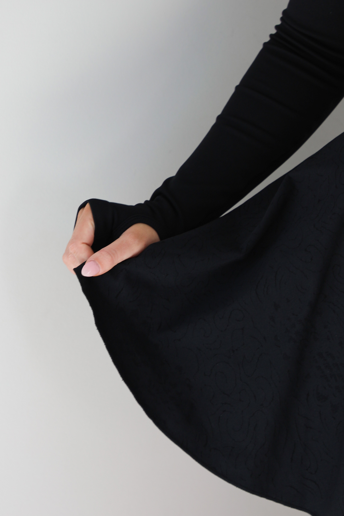 Woman wearing a black athleisure dress that has long sleeves with thumbhole cutouts, a scooped neckline, fitted bodice, and flowy skirt. The photo shows a closeup of the skirt of the dress with the squiggly textured design, and the thumbhole cut out on the sleeve. The photo is taken indoors against a white wall.