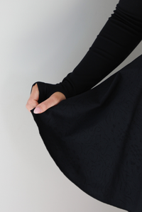 Woman wearing a black athleisure dress that has long sleeves with thumbhole cutouts, a scooped neckline, fitted bodice, and flowy skirt. The photo shows a closeup of the skirt of the dress with the squiggly textured design, and the thumbhole cut out on the sleeve. The photo is taken indoors against a white wall.