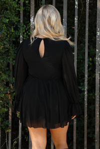 Woman model wearing a black romper that looks like a dress. The photo is taken outdoors against a gate and greenery. The photo shows the back that has a keyhole closure at the top and a cinched waistline. 