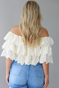 Woman wearing a cream off the shoulder, ruffled eyelet cropped top with medium wash denim shorts indoors against a white wall. The photo shows the back of the top. 