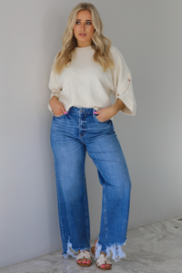 Woman wearing a cream colored quarter sleeve sweater that has ribbed details at the neckline, on the sleeves, and on the hem, with non-functional button closures on the sleeves. The sweater has a relaxed fit and is paired with a medium wash, high waisted, distressed hem denim with sand colored sandals. The photo is taken indoors against a white wall with white marble flooring. 