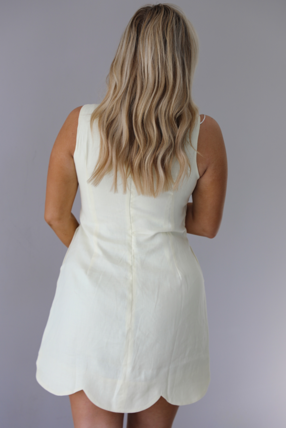 Woman wearing a pale yellow mini dress featuring a high neckline, sleeveless design, and a large scalloped hem. The photo is taken indoors against a white wall and shows the back of the dress with its zipper closure in the center. 