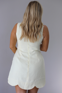 Woman wearing a pale yellow mini dress featuring a high neckline, sleeveless design, and a large scalloped hem. The photo is taken indoors against a white wall and shows the back of the dress with its zipper closure in the center. 