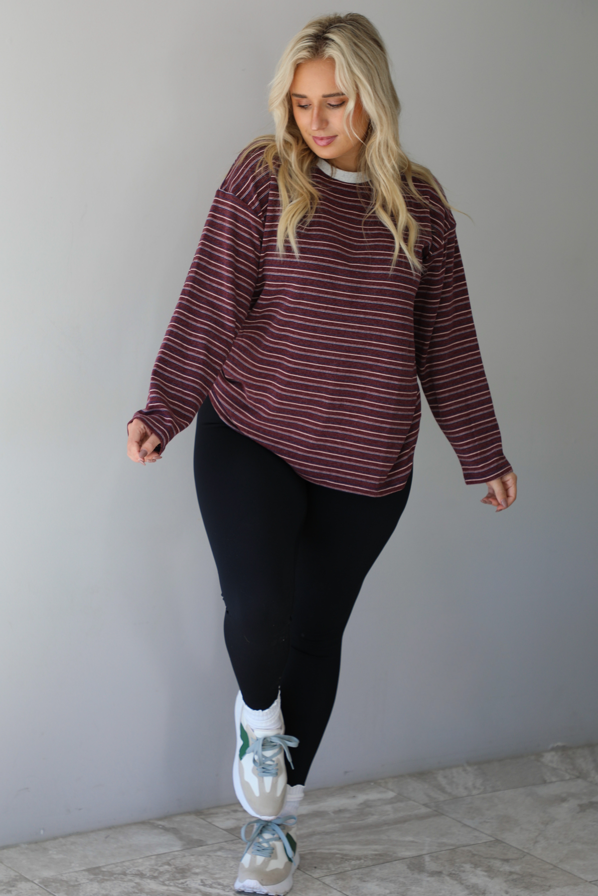 A woman wearing a burgundy striped long sleeved top paired with black leggings, high ankle socks, and sneakers, standing indoors against a white wall with white marble flooring. 