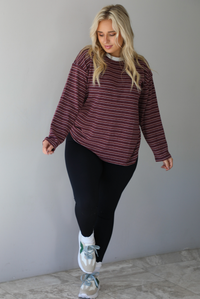 A woman wearing a burgundy striped long sleeved top paired with black leggings, high ankle socks, and sneakers, standing indoors against a white wall with white marble flooring. 
