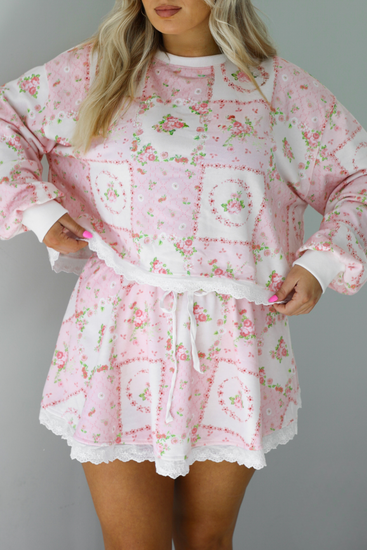 Woman wearing a two piece set featuring a pink floral set that has an oversized long sleeved top and high waisted skirt. The photo is taken indoors against a white wall.