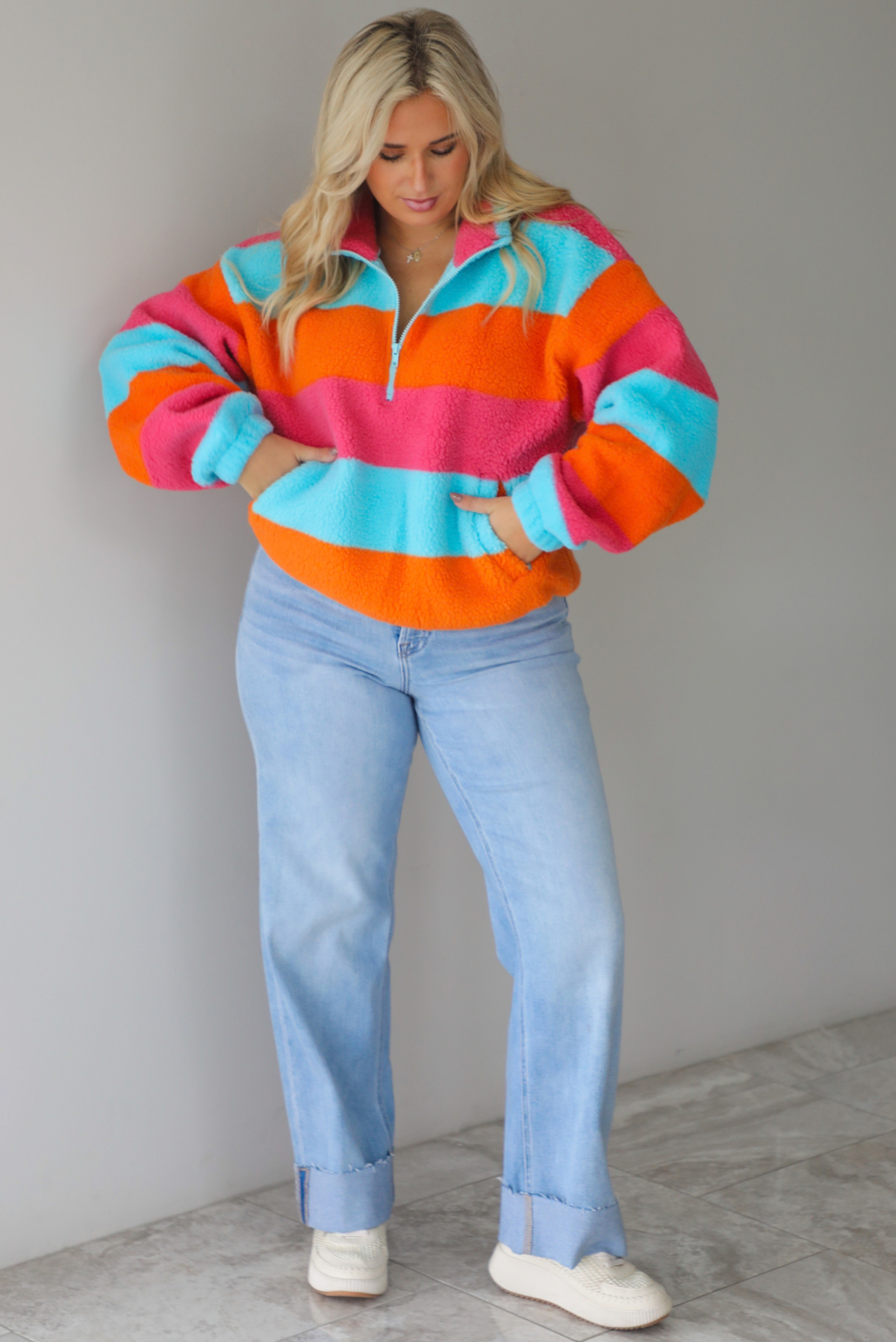 A woman wearing a turquoise, pink, and orange striped sherpa quarter-zip sweater with light wash denim jeans, standing indoors against a white wall with white marble flooring. 