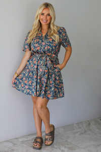 A woman modeling a short-sleeve floral mini dress with a tie waist, standing indoors against a white wall with white marble flooring. 