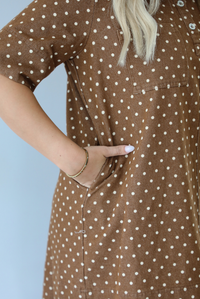 A closeup of a woman modeling a brown short-sleeve mini dress with a cream polka dot pattern, focusing on the pocket on the side of the dress, which the models' hand is resting in.