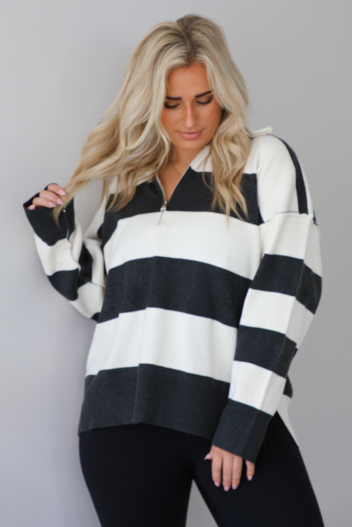 Woman wearing a charcoal and white striped quarter-zip sweater with a collared neckline, raw seam details, and a relaxed fit. It's paired with a fitted black jumpsuit. The photo is taken indoors against a white wall with white marble flooring.