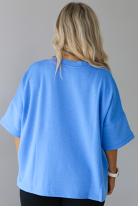 Woman wearing an oversized blue box tee with black leggings. The photo shows a closeup of the back of the tee. The photo is taken indoors against a white wall with white marble flooring. 