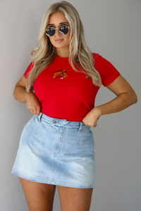Woman wearing a red fitted baby tee with an embroidered horse graphic on the front. The top is paired with a high waisted, light-wash denim skort. The photo is taken indoors against a white wall.