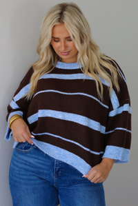 A woman wearing a brown and blue striped knit top with dark denim jeans, standing indoors against a white wall.