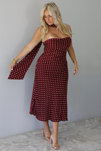 A woman wearing a strapless maroon midi dress with a white polka dot pattern with a matching scarf, standing indoors against a white wall with white marble flooring. 