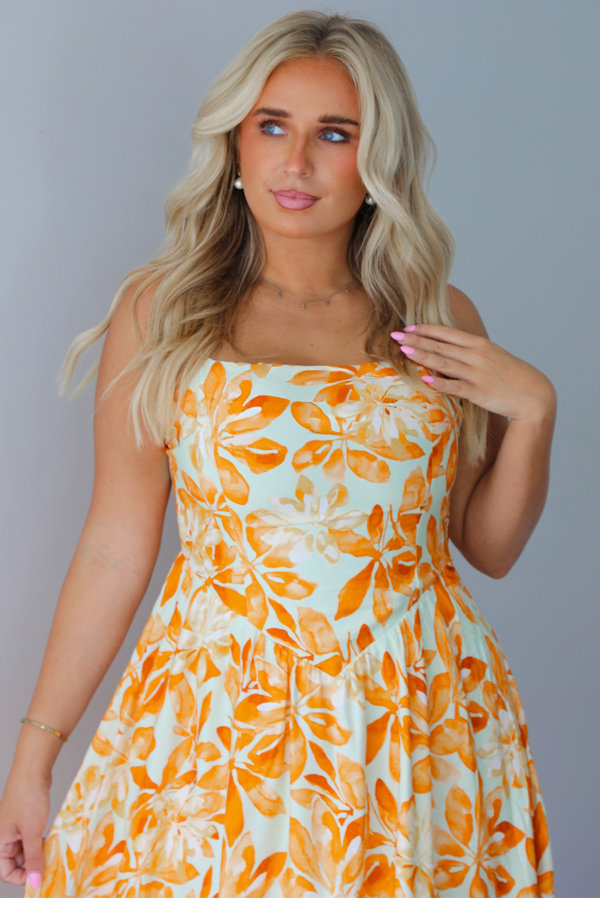 A woman modeling a spaghetti strapped, floor-length maxi dress with a white base and a vibrant marigold orange floral pattern, standing indoors against a white wall. The photo shows a closeup of the bodice of the dress, which is fitted and has a tapered V drop waist design.