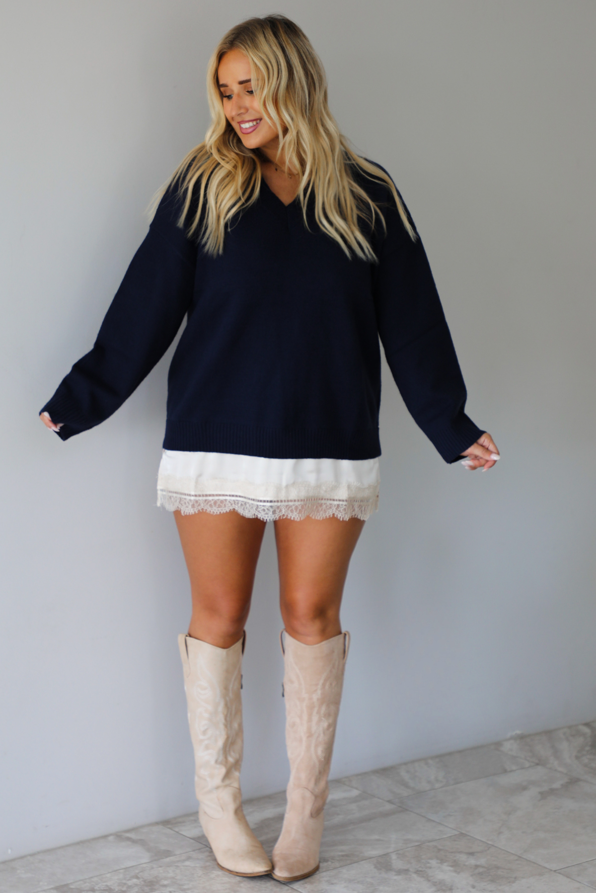 A woman wearing a navy sweater dress with a cream lace trim detail peaking out from underneath the dress giving it a layered look, paired with cream cowboy boots, standing indoors against a white wall with white marble flooring. 
