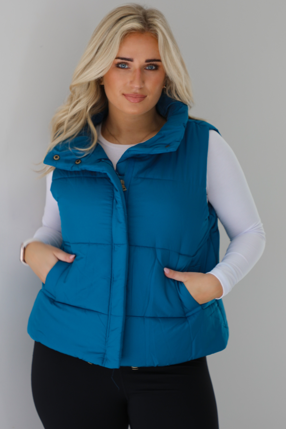 Woman wearing a teal puffer vest, white long-sleeve shirt, and black leggings indoors against a white wall. The puffer vest has a collared design, functional button closures at the neckline, a zipped closure in the front, and functional pockets on either side.