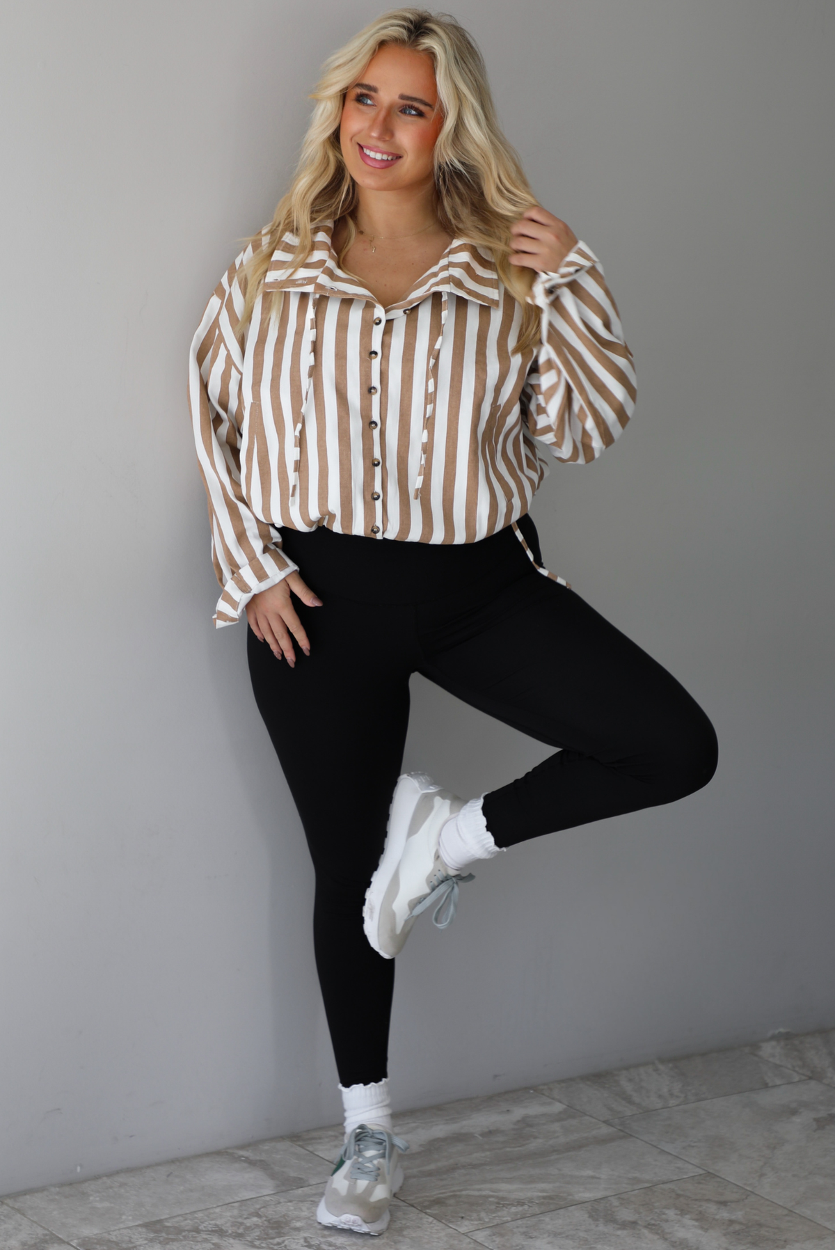 A woman wearing a tan and white striped collared long sleeved top with functional button closures along the front, worn with black leggings, white high ankle socks, and cream sneakers, standing indoors against a white wall with white marble flooring. 