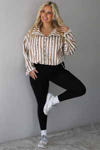A woman wearing a tan and white striped collared long sleeved top with functional button closures along the front, worn with black leggings, white high ankle socks, and cream sneakers, standing indoors against a white wall with white marble flooring. 