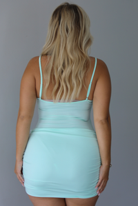 A blonde woman shown from behind modeling a light mint green spaghetti-strap mini dress with a ruched waist, standing indoors against a white wall. 
