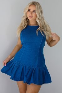 Woman wearing a dark blue denim dress that has a fitted bodice with a drop waist design thad leads to a pleated hem design. The photo shows a closeup of the dress. The photo is taken indoors against a white wall.