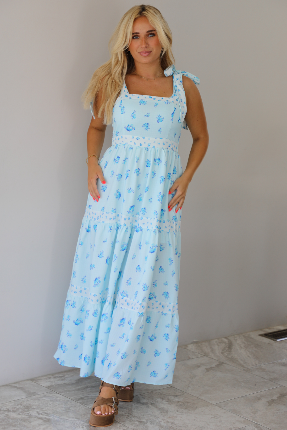Woman wearing a light blue floral maxi dress indoors against a white wall with white marble flooring. 